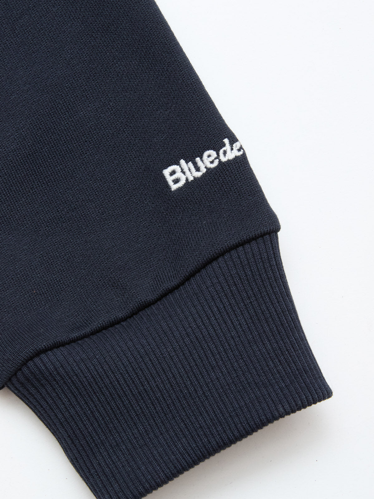 A close-up of a navy cotton jersey sleeve with ribbed cuff and partial white embroidered text reading “Bluede” near the wrist, from the Blue de Gênes BGclubhouse Rugby Sweatshirt – Navy, Clubhouse Champs Collection.