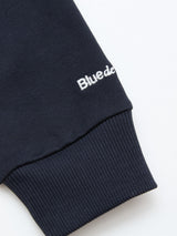 A close-up of a navy cotton jersey sleeve with ribbed cuff and partial white embroidered text reading “Bluede” near the wrist, from the Blue de Gênes BGclubhouse Rugby Sweatshirt – Navy, Clubhouse Champs Collection.