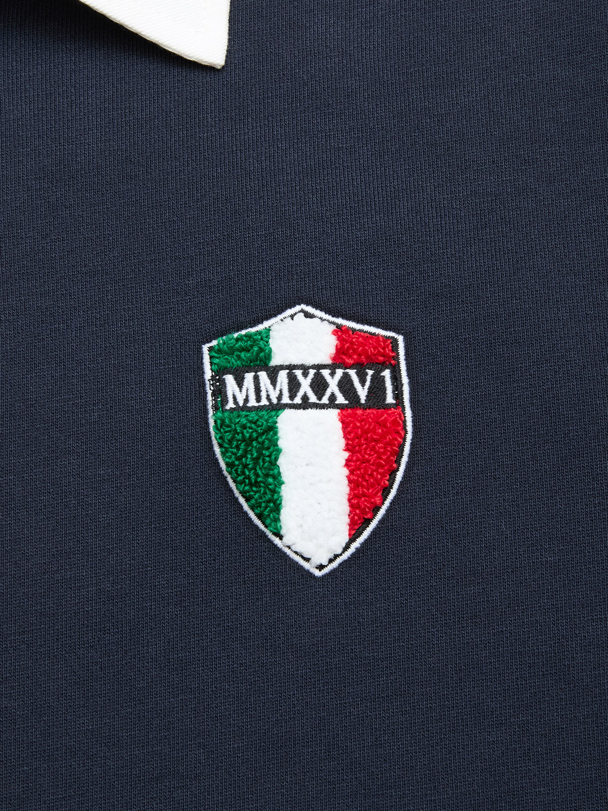 A shield-shaped patch with green, white, and red vertical stripes and “MMXXVI” in white text is sewn onto the dark cotton jersey fabric of the BGclubhouse Rugby Sweatshirt - Navy by Blue de Gênes, highlighting its athletic heritage. The collar of a white shirt is partially visible above the patch.