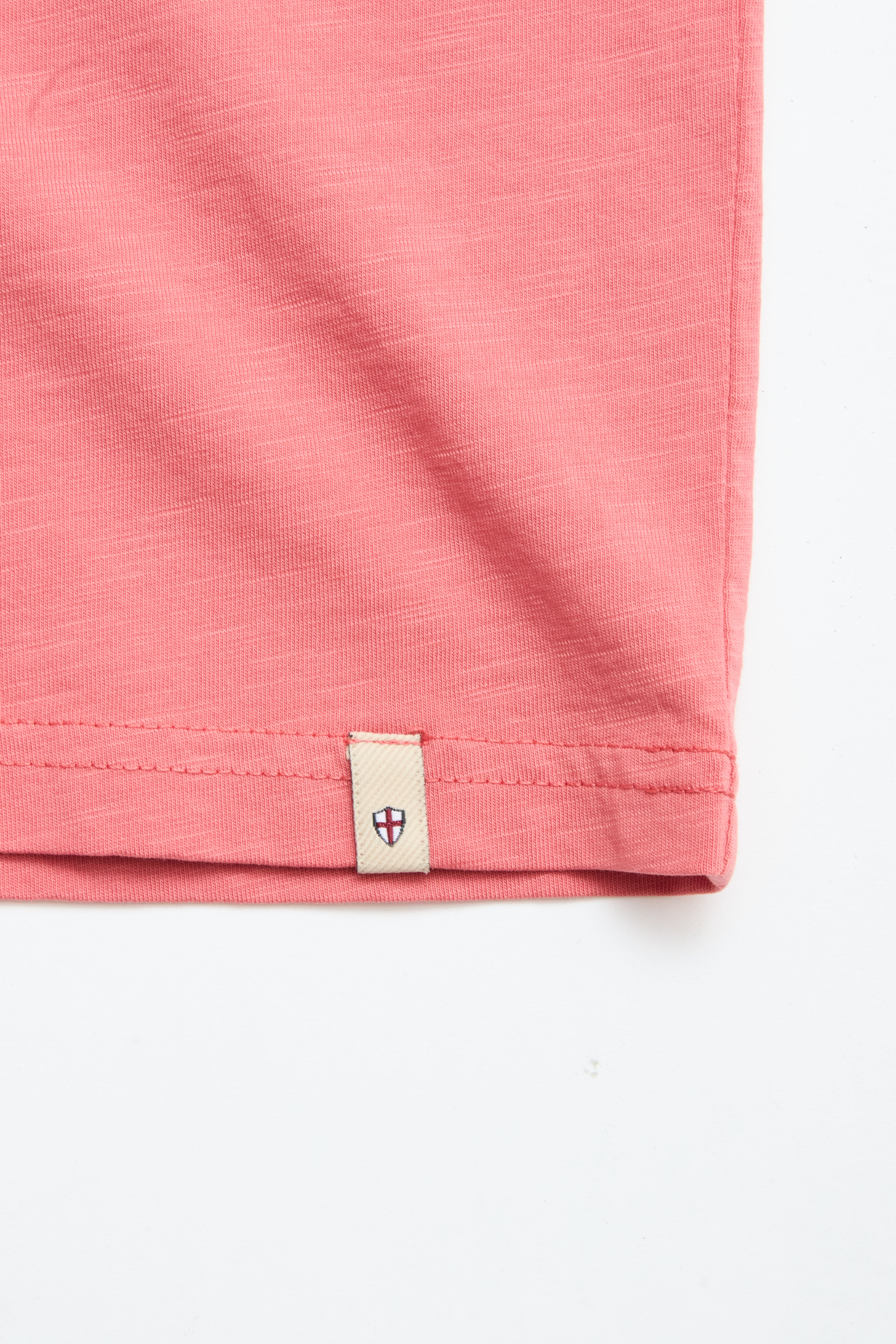 Close-up of the bottom hem of a coral BGoscar T-Shirt by Blue de Gênes in slub cotton, featuring a small beige tag with a dark shield emblem at the edge, set against a white background.
