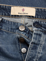 Close-up of BGvinci Striker Jeans - Dark Distress by Blue de Gênes, crafted from comfort stretch denim in blue, featuring three partly unbuttoned metallic buttons and a tan fabric label with the Blue de Gênes name and a red and white shield logo above it.