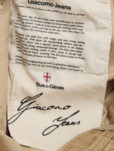 A close-up of a beige clothing label for BGgiacomo Color Jeans - Tanin by Blue de Gênes, featuring printed details about the relaxed loose fit crafted from rigid denim, with a prominent signature-style logo at the bottom.