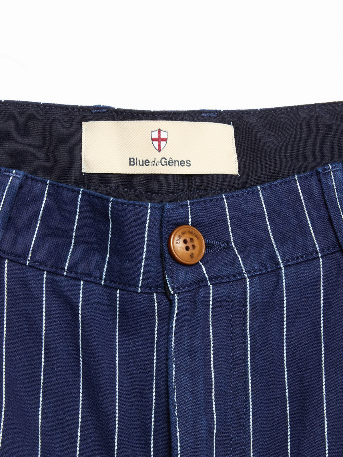 Close-up of the waistband of BGcupra Toll Shorts - Navy Stripe by Blue de Gênes, featuring a brown button and label with the brand's red and white shield logo—ideal inspiration for summer striped cotton shorts.