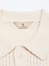 Close-up of a cream-colored BGbilly S/S Cardigan - Chalk, crafted from 100% cotton knit, featuring a buttoned collar and a fabric tag with the Blue de Gênes name below a small red cross crest, evoking subtle vintage-inspired charm.