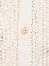 Close-up of a cream-colored, textured knit fabric with a single beige plastic button in the center, sewn onto a vertical strip of smooth fabric—showcasing the delicate lace knit pattern of the Blue de Gênes BGwalter S/S Cardigan in Chalk.