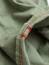Close-up of the BGpaulo Pavia Pant by Blue de Gênes in Shadow Green, featuring cotton stretch fabric, visible orange stitching, and a small red embroidered detail along the seam.