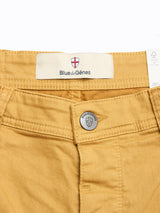 Close-up of BGpaulo Pavia Pant - Sunflower by Blue de Gênes in cotton stretch, with a metal button engraved with a shield and "Blue de Gênes," plus waistband fabric label and red and white shield logo.
