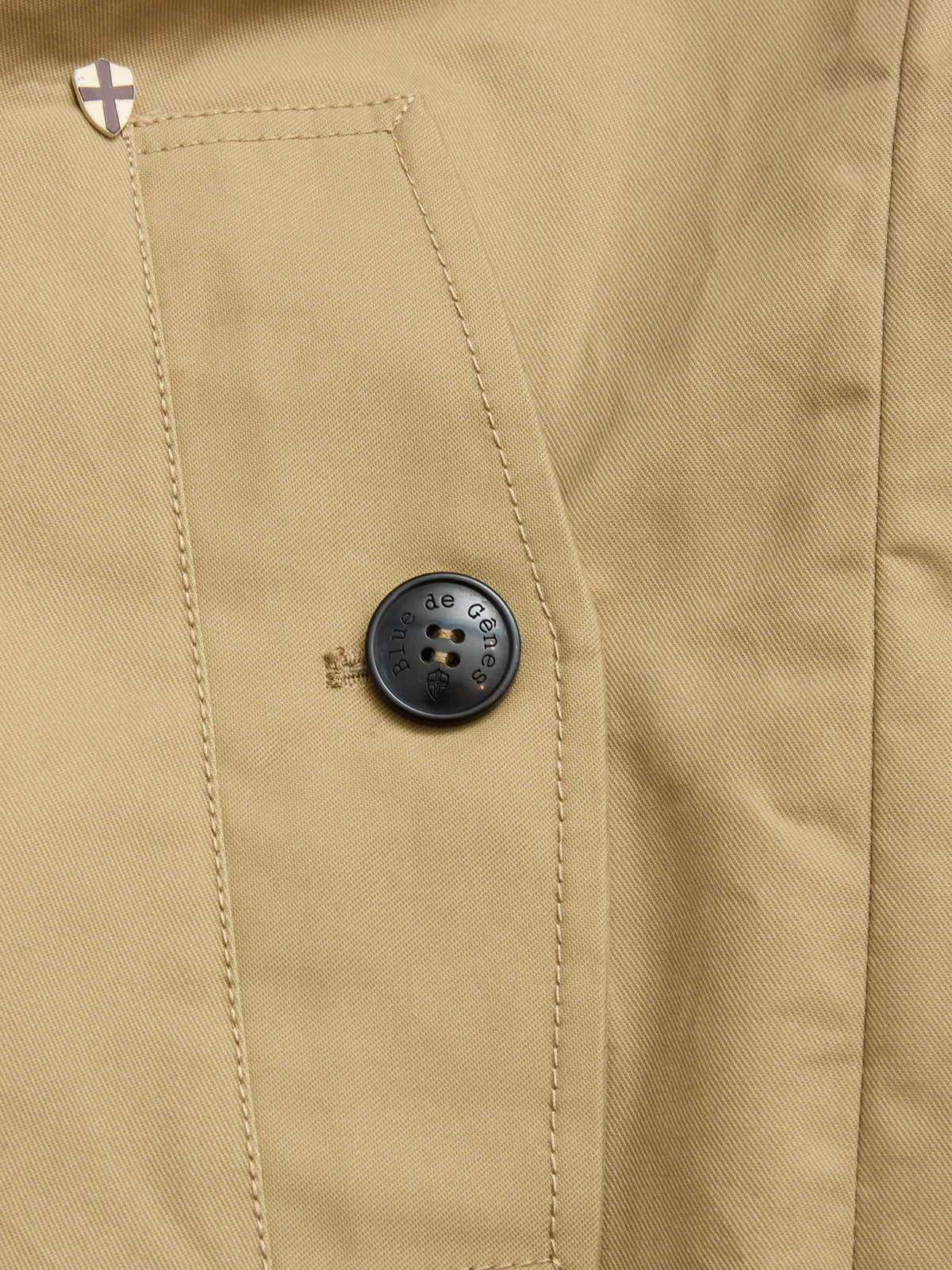 Close-up of a beige fabric pocket on the BGjacques Coat in Pale Khaki by Blue de Gênes, featuring a black engraved button and a small shield-shaped metallic emblem above the pocket.