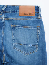 A close-up of BGvinci Hera Used Jeans by Blue de Gênes in a mid blue wash, featuring a back pocket, brown leather brand patch, orange stitching, and crafted from comfort stretch denim for easy wear.