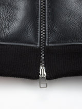 Close-up of the Blue de Gênes Rufo Shearling Jacket - Scarab, a men’s bomber jacket expertly crafted from black leather with a central silver zipper and ribbed black fabric trim at the hem—captured against a white background for a nod to timeless shearling bomber style.