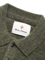 Close-up of the collar of the Malakka Polo Knit - Army, a green wool blend sweater from Blue de Gênes. A white label with the brand name “Blue de Gênes” is sewn at the neckline, topped by a red shield and cross logo. Two black corozo buttons are partially visible below the collar.