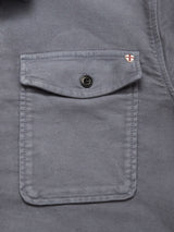 Close-up of a gray Blue de Gênes Nagano Overshirt - Dark Slate pocket with a buttoned flap and a small red and white shield patch embroidered near the top right corner, adding detail to this cotton shirt.