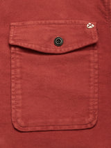 Close-up of the Nagano Overshirt - Henna by Blue de Gênes, showing a red cotton pocket with a centered black button on the flap and a small white stitched detail at the top right corner.
