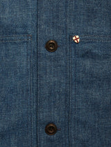 Close-up of the Blue de Gênes Yuma Fano Jacket in denim blue—features heavy cotton blend fabric, two black buttons, visible stitching, and a small shield-shaped pin with a red cross on white above the right pocket.