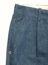 Close-up of Blue de Gênes Shogun Fano Pant in Denim Blue, highlighting the heavy cotton blend, waistband, belt loops, pleats, and a small metallic pin with a white background and black cross with gold details near the pocket.