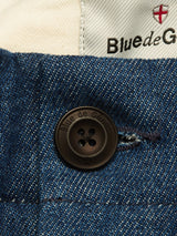 Close-up of denim blue fabric and a black Blue de Gênes button on the Shogun Fano Pant. Above the button, part of a label featuring the brand name and red cross logo appears on the white inner lining, reflecting their contemporary style.