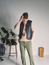 A man in a brown beanie, black vest, and green pants stands indoors by a potted plant, holding a camera. His black vest is the Saga Shearling Gilet - Scarab by Blue de Gênes, a stylish reversible gilet crafted from German Merino sheepskin.