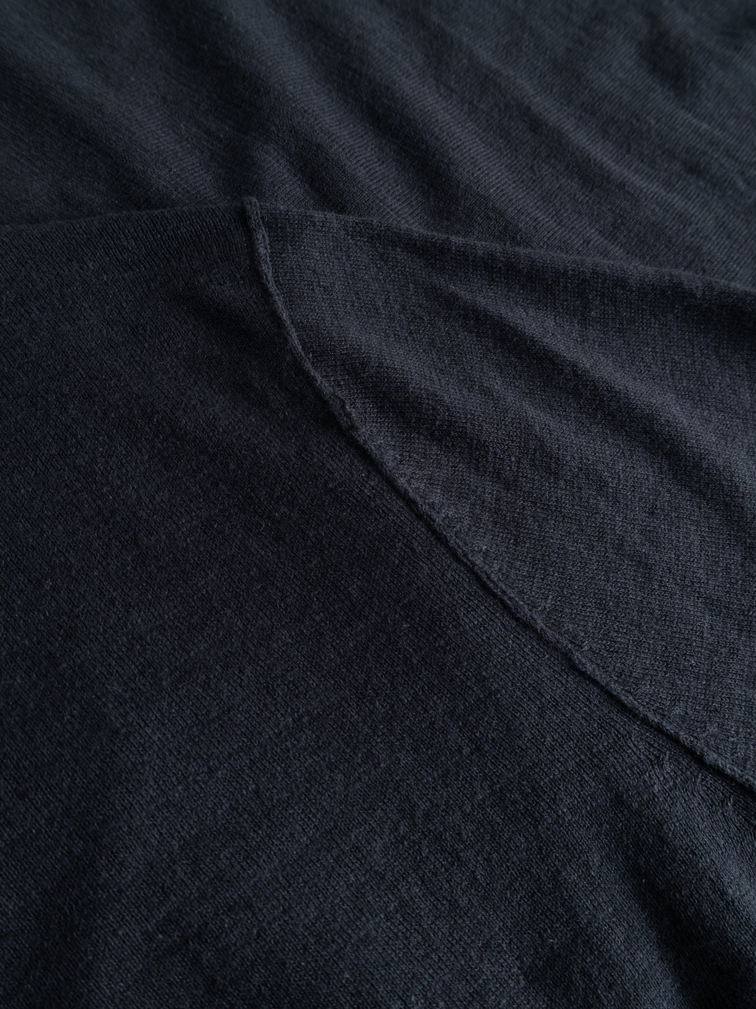 A close-up of Blue de Gênes' BGmonty Knit in Dark Navy shows soft, dark textured fabric with visible stitching and a diagonal roll-rib hem, featuring a slight sheen.