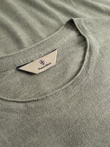 A close-up of the BGmonty Knit in shadow green, focusing on the crewneck collar and a beige label featuring the Blue de Gênes shield logo and branding. The fabric texture and stitching of this regular fit piece are clearly visible, highlighting its quality cotton linen blend.