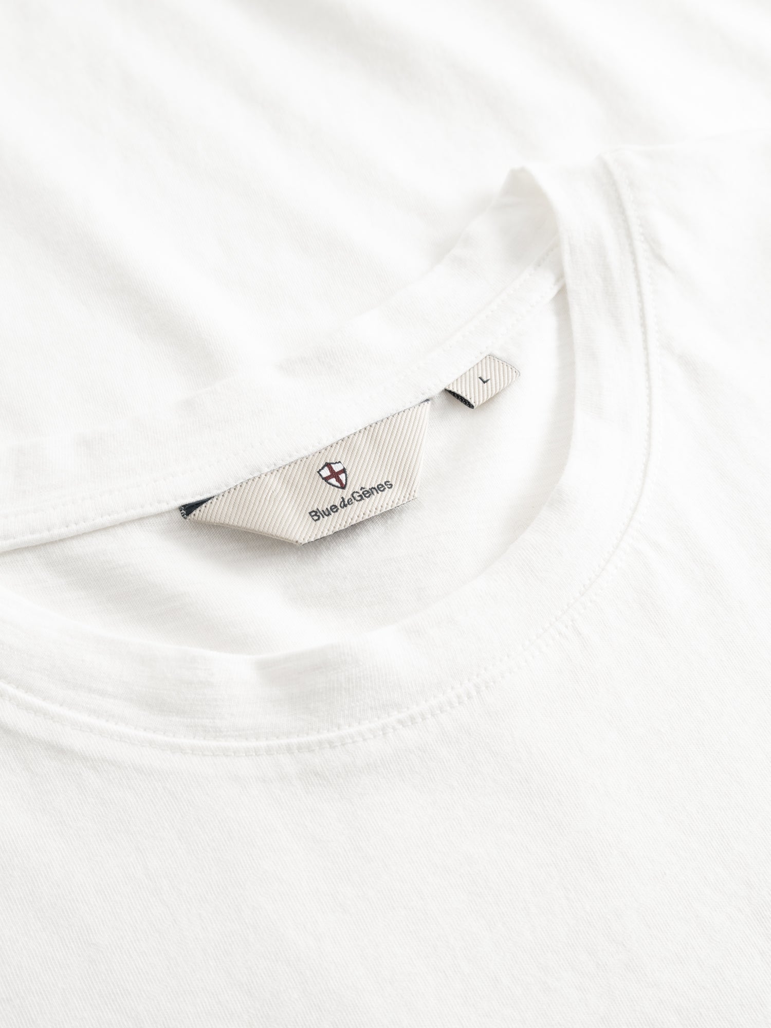 Close-up of a white Blue de Gênes BGoscar T-Shirt - Snow Fall, crafted from 100% cotton, featuring a visible beige label inside the collar with logo and “Save & Corona” text.