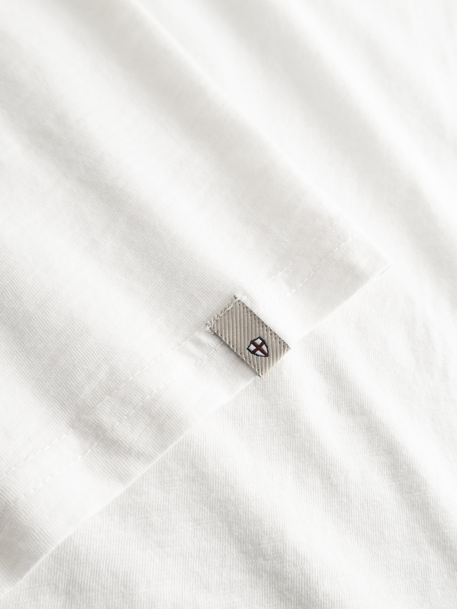 Close-up of the BGoscar T-Shirt - Snow Fall by Blue de Gênes, a soft 100% cotton crewneck in white with a small gray geometric logo tag stitched near the hem and neat visible edge stitching.