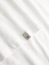 Close-up of the BGoscar T-Shirt - Snow Fall by Blue de Gênes, a soft 100% cotton crewneck in white with a small gray geometric logo tag stitched near the hem and neat visible edge stitching.