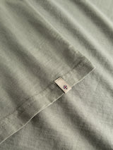 A close-up of the BGoscar T-Shirt - Shadow Green by Blue de Gênes shows the light gray slub cotton fabric, stitched hem, a small tag with a red and blue symbol, and the soft crewneck texture.