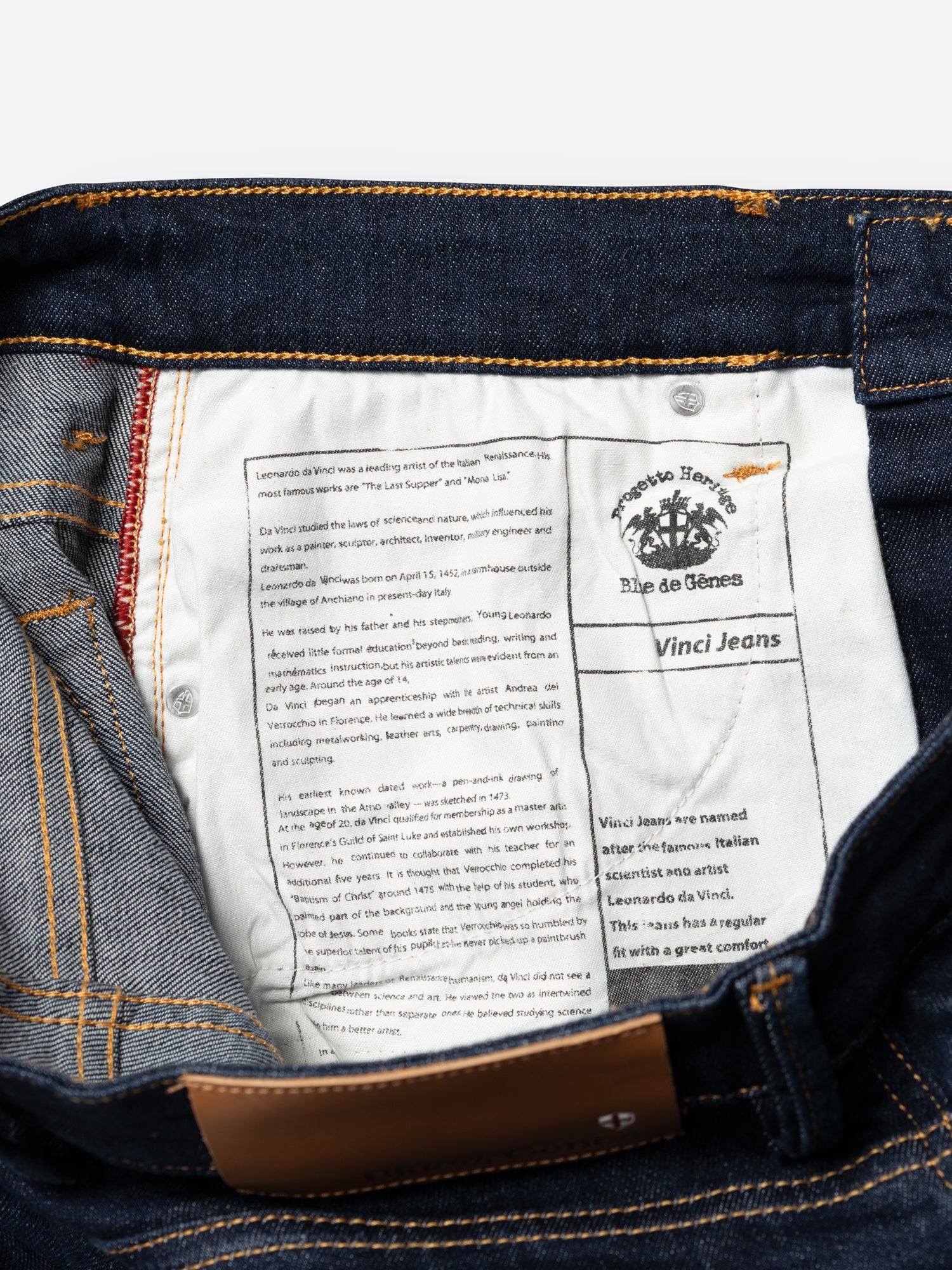 Close-up of BGvinci Pala Dark V2 Jeans by Blue de Gênes, featuring dark blue denim, a white fabric label with brand and heritage details, comfort stretch, yellow stitching, and part of a brown leather patch.