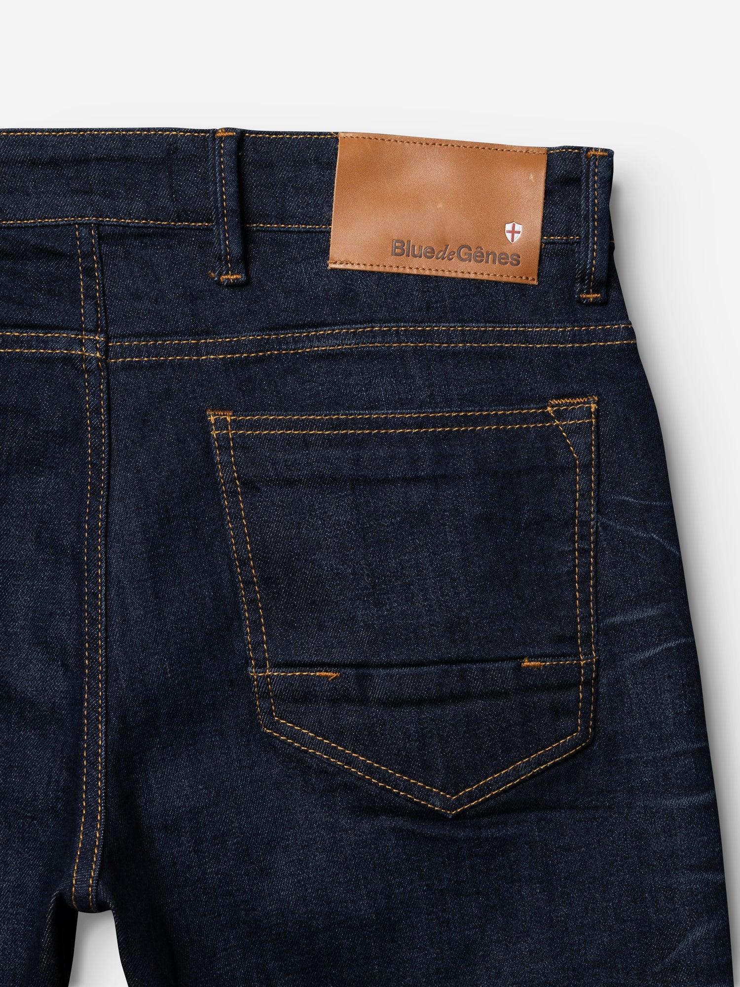 Close-up of the BGvinci Pala Dark V2 Jeans by Blue de Gênes in dark blue denim, showing orange stitching, comfort stretch fabric, a back pocket, and a tan leather waistband patch with the brand name and shield logo.