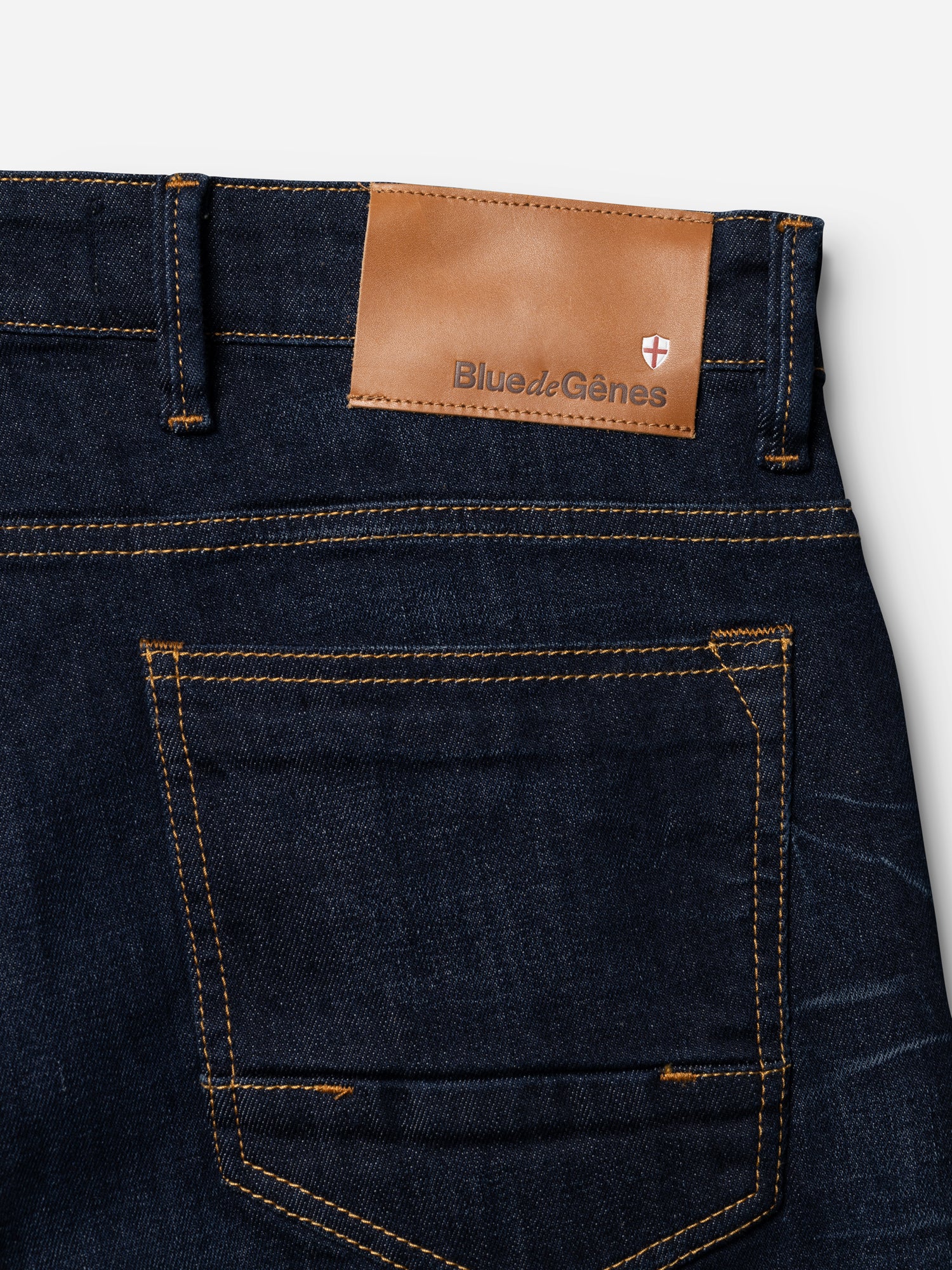 Close-up of the back pocket on BGvinci Pala Dark V2 Jeans in dark blue denim by Blue de Gênes, featuring orange stitching and a brown leather patch with brand name and a red and white emblem.