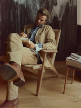 A man with a beard, dressed in Blue de Gênes BGmenzo Rory Chino - Khaki pants and a matching blazer, sits in a wooden chair writing in a notebook. He pairs his outfit with brown boots. Behind him is a dark abstract wall, and nearby stands a small table stacked with books.