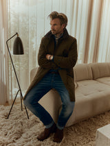 A man wearing the Blue de Gênes BGjacques AW Coat in brown, paired with jeans and brown boots, leans against a beige sofa in a stylish living room featuring sheer curtains, a modern floor lamp, and a shaggy rug.