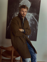 A man with blonde hair and a beard sits on a wooden chair, arms crossed, wearing the Blue de Gênes BGjacques AW Coat in brown—crafted from heavy cotton—in front of abstract black-and-white artwork.