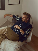 A man with tattoos lounges on a modern chair with a fur throw, wearing a striped shirt, tan pants, a watch, and the Blue de Gênes BGdand Waistcoat in dark navy, in a cozy room decorated with framed pictures on the wall.