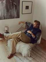 A man with blond hair relaxes on a modern chair draped with a furry throw, wearing a striped shirt, vest, and the BGliam Nebraska Chino - Brown by Blue de Gênes, paired with brown boots. Artwork and a lamp decorate the cozy, minimal room.