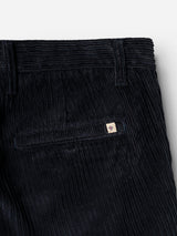 Close-up of navy stretch corduroy pants from Blue de Gênes, the BGpalco Cord Chino, showing a back pocket with a small, rectangular logo patch featuring a red and white shield. The fabric displays the distinct vertical ribbed texture characteristic of these relaxed fit corduroy trousers.