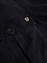 Close-up of a navy fabric pocket on the Blue de Gênes BGbarnaba Iro Overshirt, featuring a black button and a small gold shield-shaped pin with a black cross, crafted from a lyocell-cotton blend.