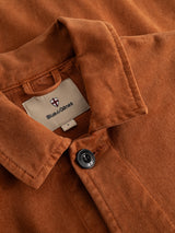 Close-up of the BGbarnaba Iro Overshirt in Ginger Bread Orange by Blue de Gênes—a workwear-inspired, rust-hued collared overshirt featuring a single black button and a visible fabric tag displaying the Blue de Gênes shield logo above the brand name. Crafted from soft comfort fabric, this piece embodies rugged style with everyday versatility.