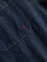 Close-up of the BGgregor Denim Shirt - Denim Blue by Blue de Gênes, showcasing a blue indigo denim pocket with orange stitching. A small red and white X is embroidered near the top center of the pocket. The shirt features a subtle texture and visible weave, highlighting its classic craftsmanship.