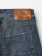 Here’s your requested rewrite using the provided product data: Close-up of the BGgiacomo Alfie Jeans in mid blue denim by Blue de Gênes, showcasing a brown leather patch embossed with the brand’s name and crest above the back pocket. These jeans are crafted from comfort stretch denim, featuring visible stitching and faded detailing for an effortlessly relaxed look.