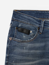 Close-up of the Blue de Gênes BGvinci OM Jeans - Denim wash, highlighting the waistband, front pocket, belt loop, and a distinctive small red and white shield-shaped patch on the coin pocket. The authentic worn-in denim features a faded and gently distressed texture. Classic five-pocket styling enhances the timeless appeal of these jeans.