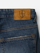 Close-up of the back pocket on the BGrepi Presley Jeans - Denim Blue by Blue de Gênes, featuring a brown leather patch with an embossed shield emblem near the waistband. The comfort stretch denim fabric showcases visible contrast stitching.