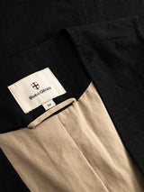 Close-up of the BGduncan Ian Gilet in black by Blue de Gênes, featuring a beige lining and a visible clothing label with the brand’s shield logo above it. This refined piece, part of a three-piece suit, is neatly laid out to showcase its lightweight textured fabric.