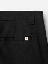 Close-up of the BGpalco Ian Pant by Blue de Gênes in black linen-wool. A welt pocket is accented with a small white tag bearing a red shield emblem at the top edge. The richly textured herringbone fabric and tapered silhouette create a modern, refined look.