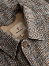 Close-up of the BGmungo Check Overshirt by Blue de Gênes, showcasing its grey check herringbone pattern crafted from a wool-blend. The design features a buttoned collar and an inner label reading Blue de Gênes, topped with a small red and white shield logo. This men’s regular fit piece merges classic style with modern comfort.