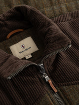 Close-up of the Blue de Gênes BGalec Skotch Waistcoat in brown check, featuring visible zipper and corduroy panels, with a fabric label reading “Blue de Gênes L.” The waistcoat is laid on top of a textured, checked wool fabric background.