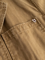 Close-up of the BGgabbiano Rory Blazer - Khaki by Blue de Gênes, made from tan washed herringbone fabric and featuring a front pocket. A small enamel pin with a shield and black cross is attached to the pocket, highlighting the relaxed blazer’s unconstructed design.