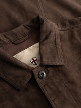 Close-up of the BGvalan Suede Jacket - Dark Brown by Blue de Gênes, focusing on the collar and button, with a beige tag featuring a partially visible red-and-black shield emblem peeking out from beneath the collar.