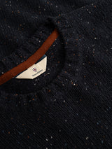 A close-up of the BGadamo Crewneck Knit in Dark Navy by Blue de Gênes, showcasing its dark blue speckled wool knit with multicoloured nep threads that add subtle pops of color throughout. The ribbed crewneck collar features a partially visible white tag inside the neckline, adorned with Blue de Gênes’ shield logo and text.