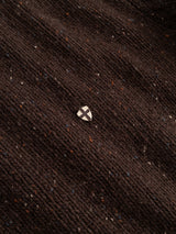 A small shield-shaped pin featuring a black cross on a white background is placed on the BGadamo Crewneck Knit - Hot Fudge Brown by Blue de Gênes, its textured wool fabric in rich brown flecked with multicoloured nep.